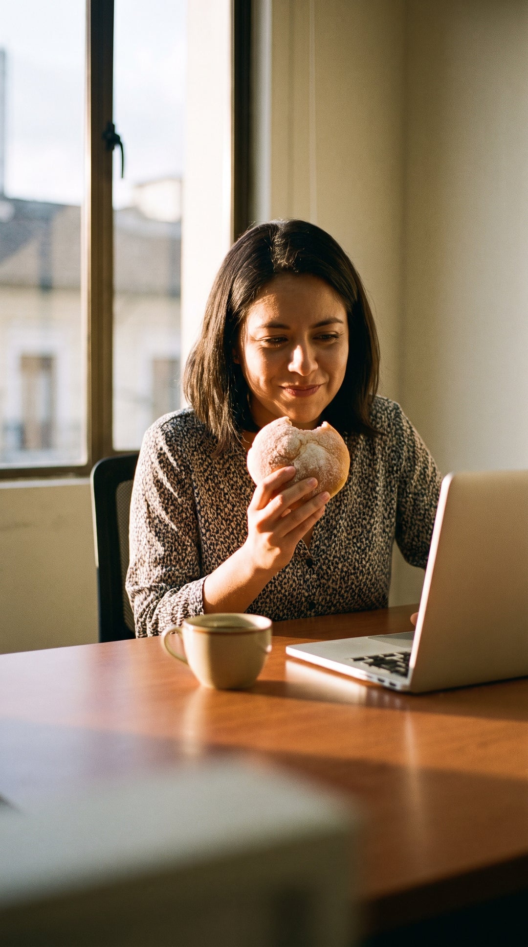 Mujer en su escritorio a media tarde, con una taza de café y un pan dulce a medio comer